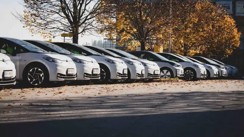 Reihe von mehreren weißen E-Autos, die auf einem Parkplatz im Herbst unter bunten Bäumen nebeneinander stehen.