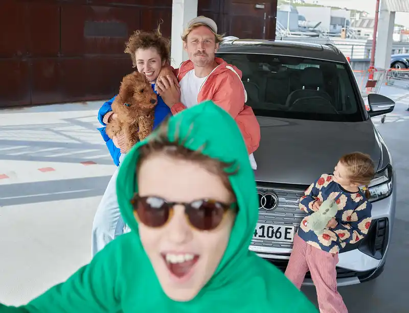 Eine Familie steht mit einem Hund vor einem geparkten E-Auto, während ein Kind im Vordergrund fröhlich in die Kamera lacht.