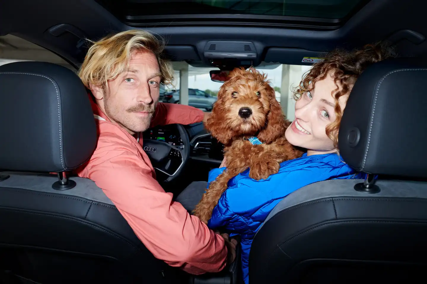 Ein Paar sitzt im Innenraum eines E-Autos und hält einen kleinen Hund auf dem Schoß, während beide lächelnd in die Kamera schauen.
