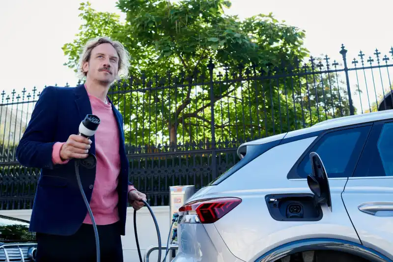 Mann steht neben einem weißen E-Auto und hält ein Ladekabel an einer Ladesäule.