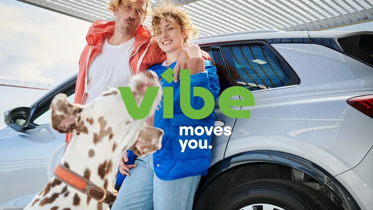Ein Paar lehnt lächelnd an einem silbernen E-Auto, während ein Dalmatiner im Vordergrund nach oben schaut und das Logo von vibe eingeblendet wird.