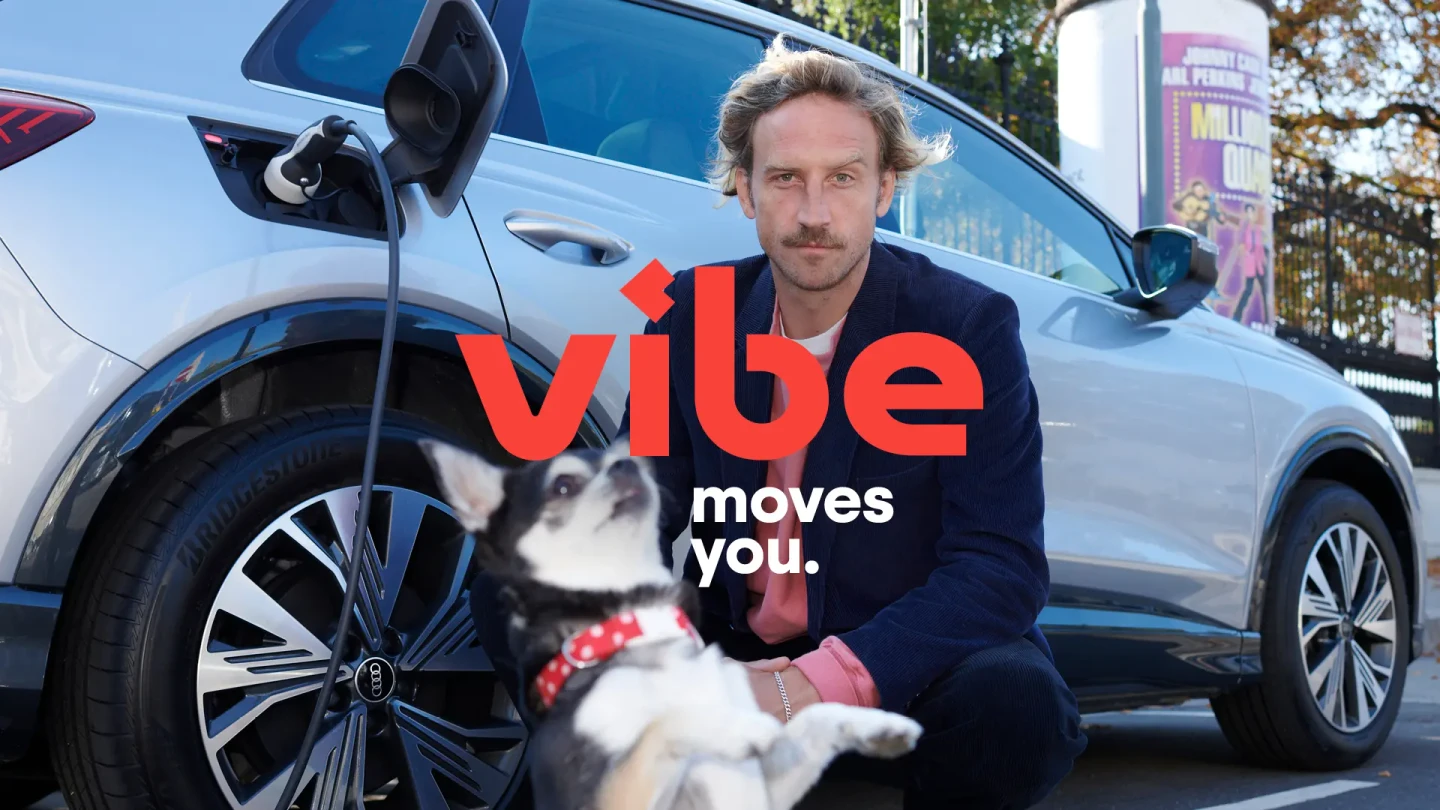Ein Mann sitzt vor einem silbernen E-Auto mit angeschlossenem Ladekabel, während ein kleiner Hund im Vordergrund zu sehen ist und das Logo von vibe eingeblendet wird.