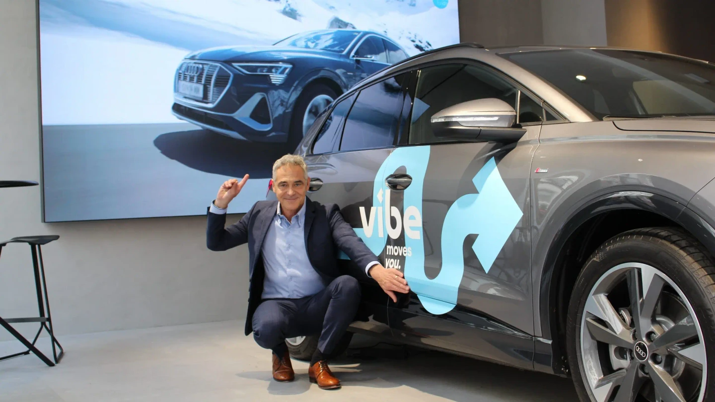 Christian Clerici von vibe kniet neben einem gebrandeten E-Auto und zeigt lächelnd auf ein großes Bild eines Audi E-Autos.
