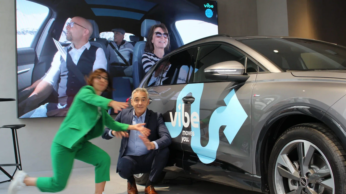 Christian Clerici und Katharina Wirth-Gorbach, Head of Brand, PR & Partnerships bei vibe, posieren neben einem gebrandeten E-Auto vor einer großen Leinwand mit einer Fahrszene im Hintergrund.