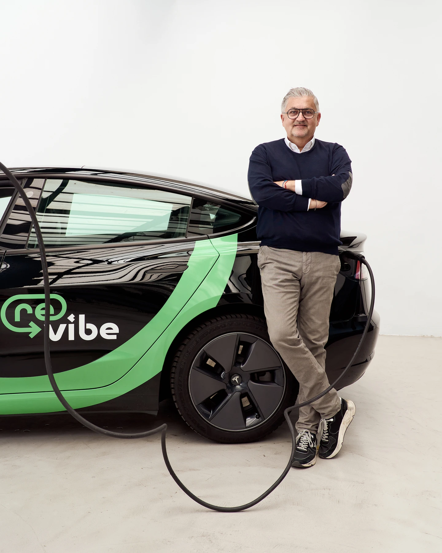 vibe CEO Martin Rada steht mit verschränkten Armen neben einem schwarzen revibe E-Auto in einem Studio, während ein Ladekabel am Fahrzeug angeschlossen ist.