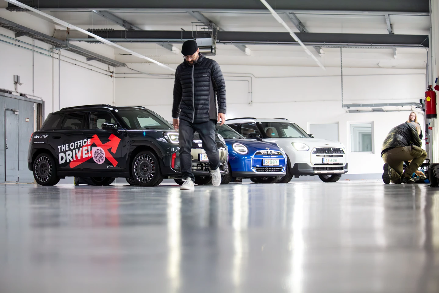 Aufnahme in einer hellen Industriehalle: Mehrere E-Autos stehen nebeneinander geparkt, im Vordergrund geht eine Person über den glänzenden Hallenboden, während im Hintergrund weiteres Produktionsequipment und Crewmitglieder zu sehen sind.