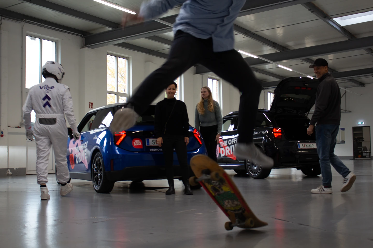 Szene in einer hellen Industriehalle: Mehrere Personen aus dem vibe Team bewegen sich zwischen E-Autos mit „The Office Drive!“ Branding. Ein Mann fährt auf einem Skateboard, während im Hintergrund ein maskierter vibe Markencharakter und weitere Teammitglieder an den Fahrzeugen stehen.