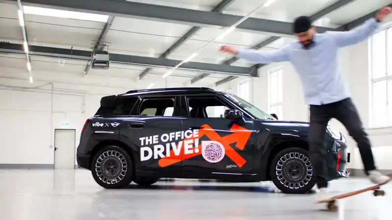 Ein schwarzes E-Auto mit „The Office Drive!“ und vibe Branding steht in einer hellen Industriehalle. Im Vordergrund springt ein Mann auf einem Skateboard vorbei, was der Szene eine dynamische, spielerische Stimmung verleiht.