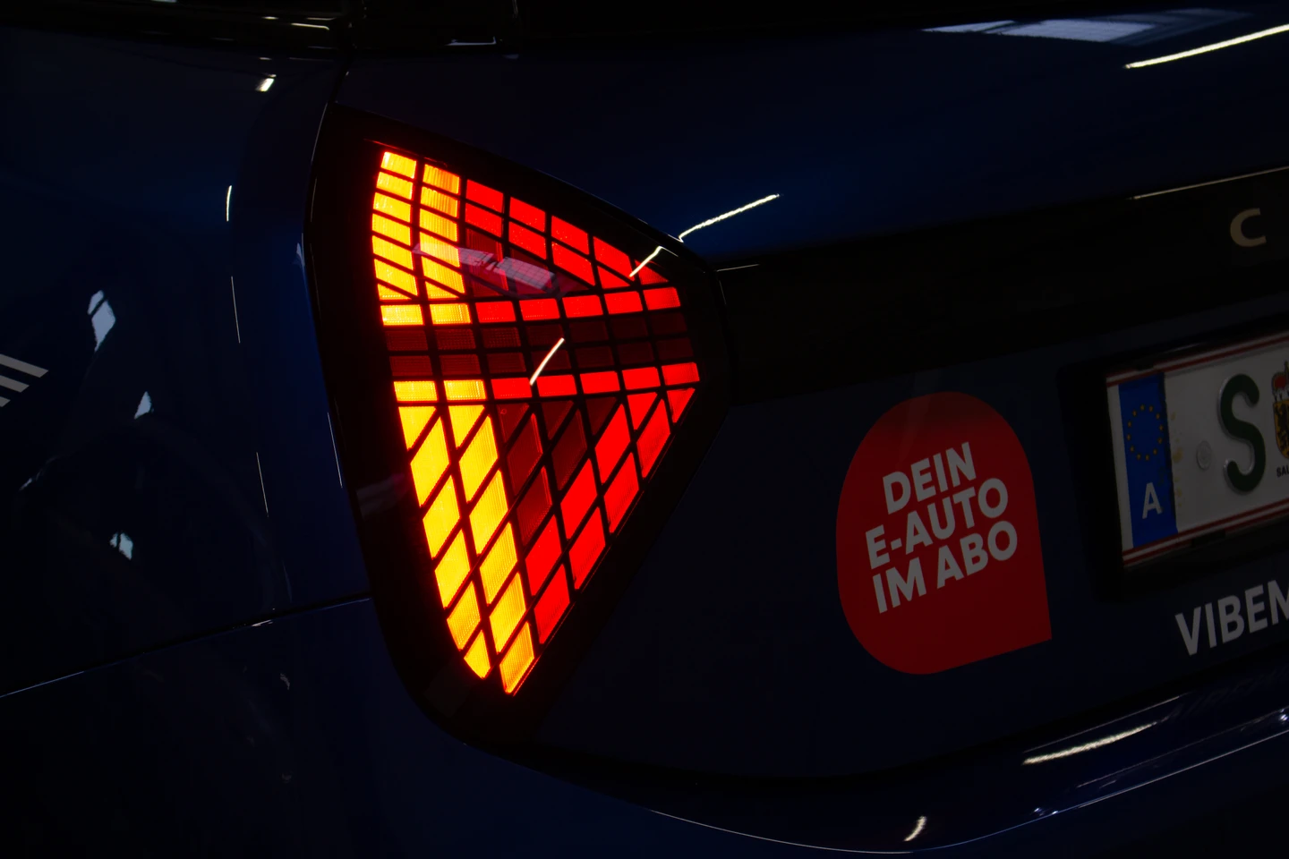 Detailaufnahme des Rücklichts eines dunklen E-Autos in einer Halle. Neben dem leuchtenden, pfeilförmigen Rücklicht ist ein Aufkleber mit der Aufschrift „Dein E-Auto im Abo“ sowie ein Kennzeichen mit vibe Branding zu sehen.