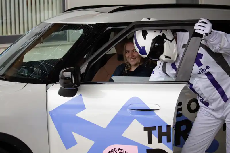 Eine Frau von spl tele und electrify sitzt lächelnd auf dem Fahrersitz eines E-Autos, während eine Person im weißen vibe-Anzug mit Helm sich durch die geöffnete Fahrertür ins Fahrzeug lehnt. Auf der Autoseite ist das Branding von „The Office Drive!“ zu sehen.