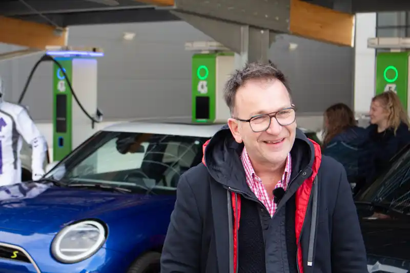Thomas Robitza, CTO der electrify GmbH, steht lächelnd vor einem E-Auto an einer Ladeinfrastruktur mit mehreren Ladesäulen, während im Hintergrund weitere Teammitglieder zu sehen sind.