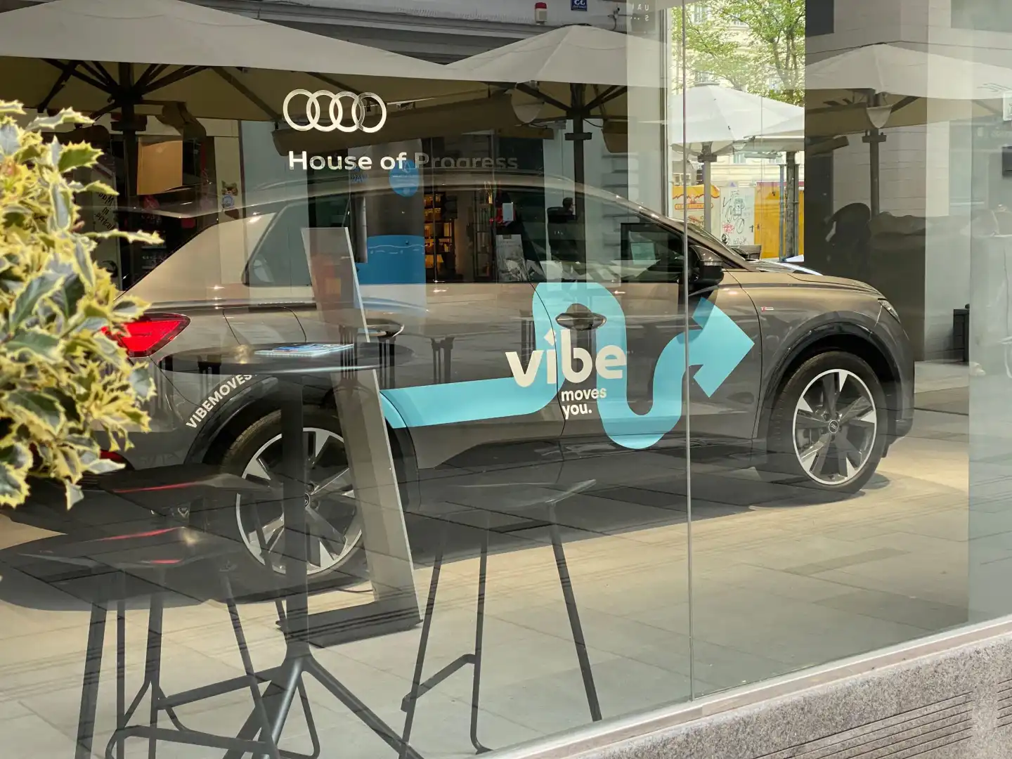 Ein gebrandetes vibe E-Auto steht vor dem Audi House of Progress und ist durch eine Glasfront zu sehen. Das Fahrzeug ist seitlich abgebildet und trägt das Logo „vibe moves you“, im Vordergrund spiegeln sich Stühle und Pflanzen im Glas.