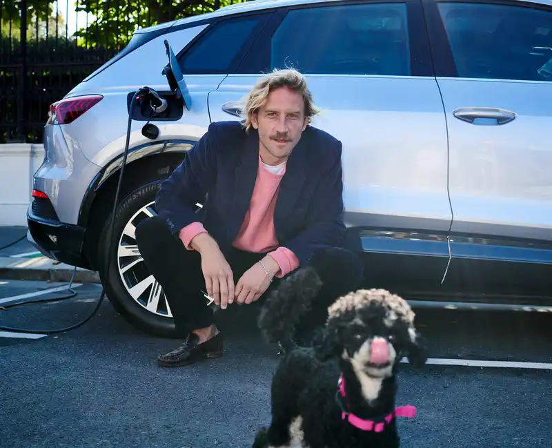 Mann hockt neben einem weißen E-Auto an einer Ladestation, im Vordergrund steht ein Hund.