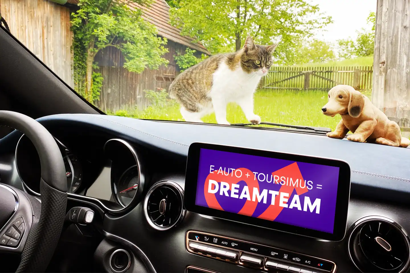 Blick aus dem Innenraum eines E-Autos: Auf dem Armaturenbrett steht eine kleine Hundefigur, draußen läuft eine Katze vor der Windschutzscheibe über eine grüne Wiese. Auf dem Display im Auto steht der Schriftzug „E-Auto + Tourismus = Dreamteam“.